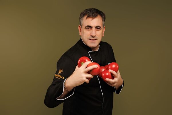 A chef in a black uniform holds several ripe red tomatoes in both hands while standing against a plain olive-green background.