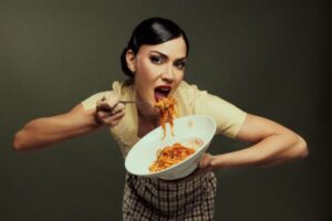 A woman holding a bowl of spaghetti in one hand and lifting a forkful toward her mouth with an expressive, playful pose against a plain dark background.