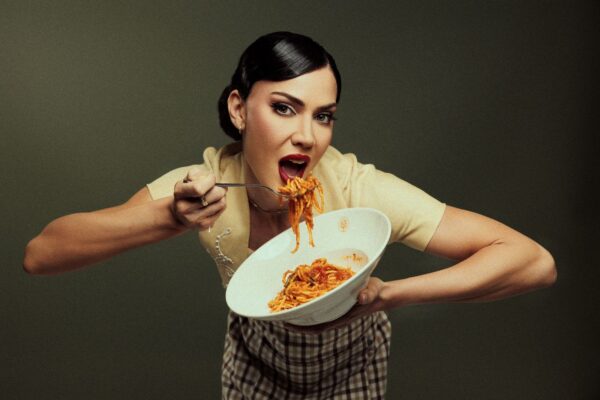 A woman holding a bowl of spaghetti in one hand and lifting a forkful toward her mouth with an expressive, playful pose against a plain dark background.