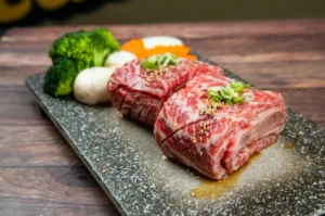 Close-up of thick, marbled cuts of raw beef arranged on a stone platter, garnished with sliced green onions and sesame seeds. Fresh vegetables, including broccoli, mushrooms, and carrot slices, are placed beside the meat.