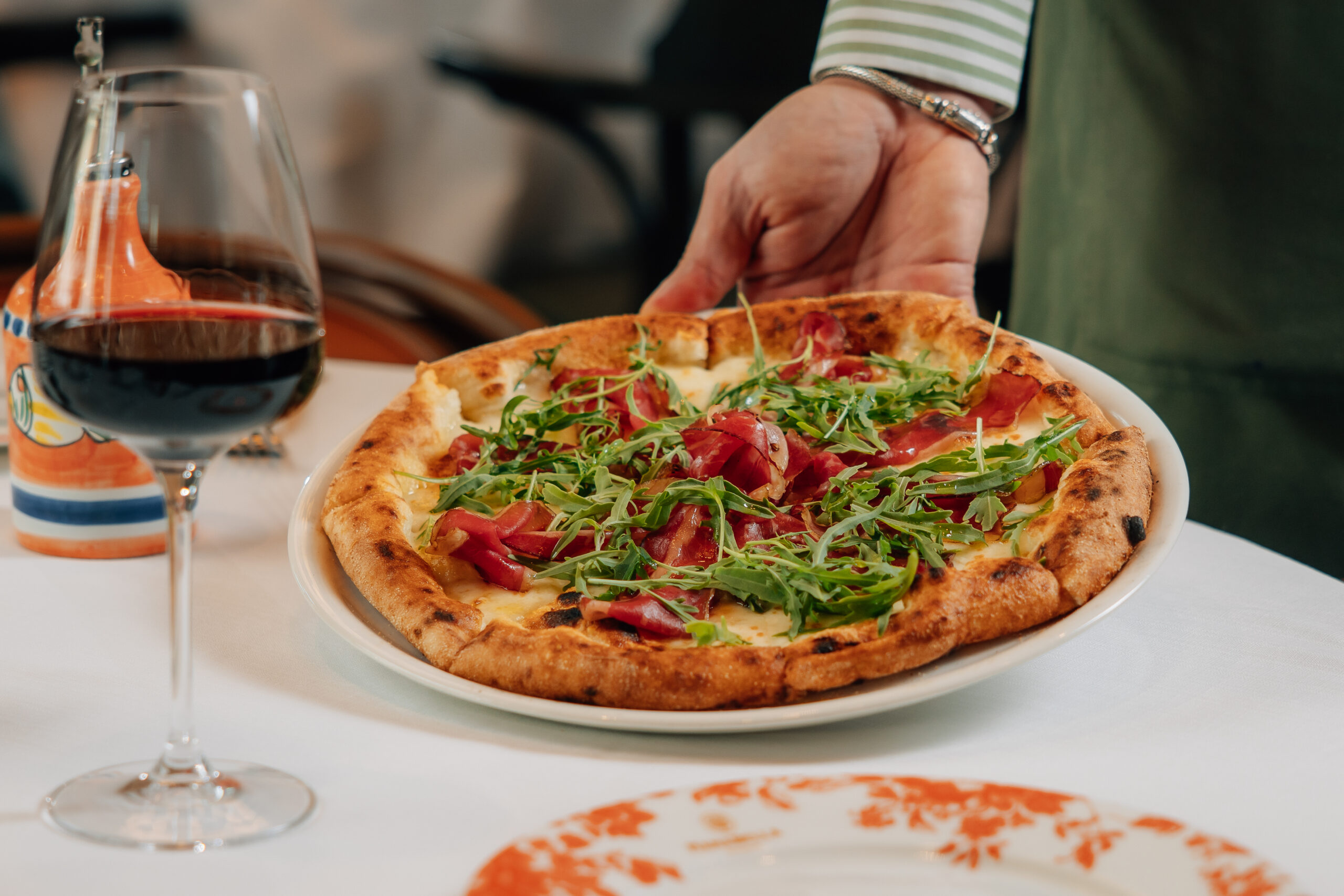 Fresh Italian pizza topped with cured meat and rocket, served at Mamabella with a glass of red wine.