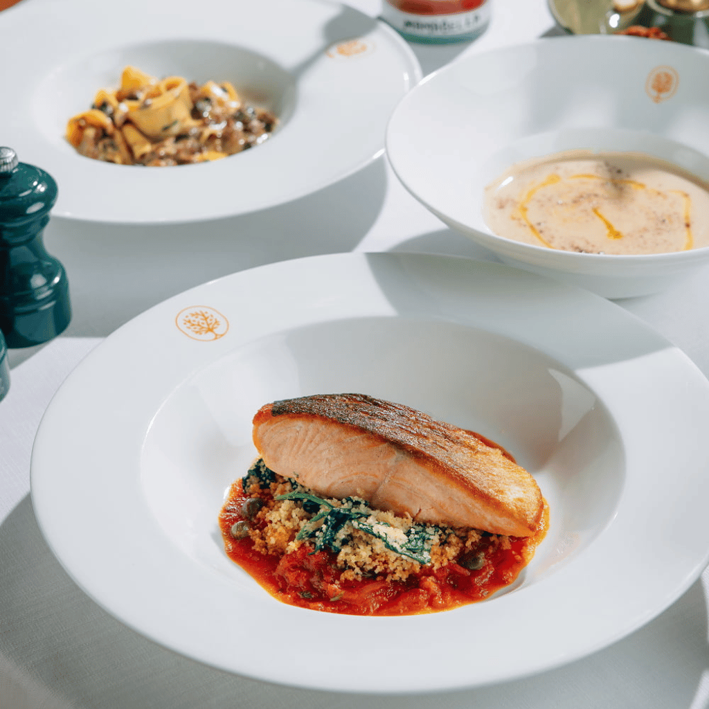 Grilled salmon served with tomato sauce, greens and breadcrumbs at Mamabella, with pasta and soup in the background.