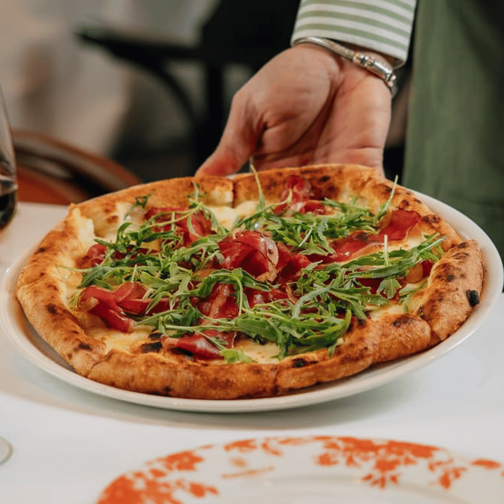 Fresh Italian pizza topped with cured meat and rocket, served at Mamabella with a glass of red wine.